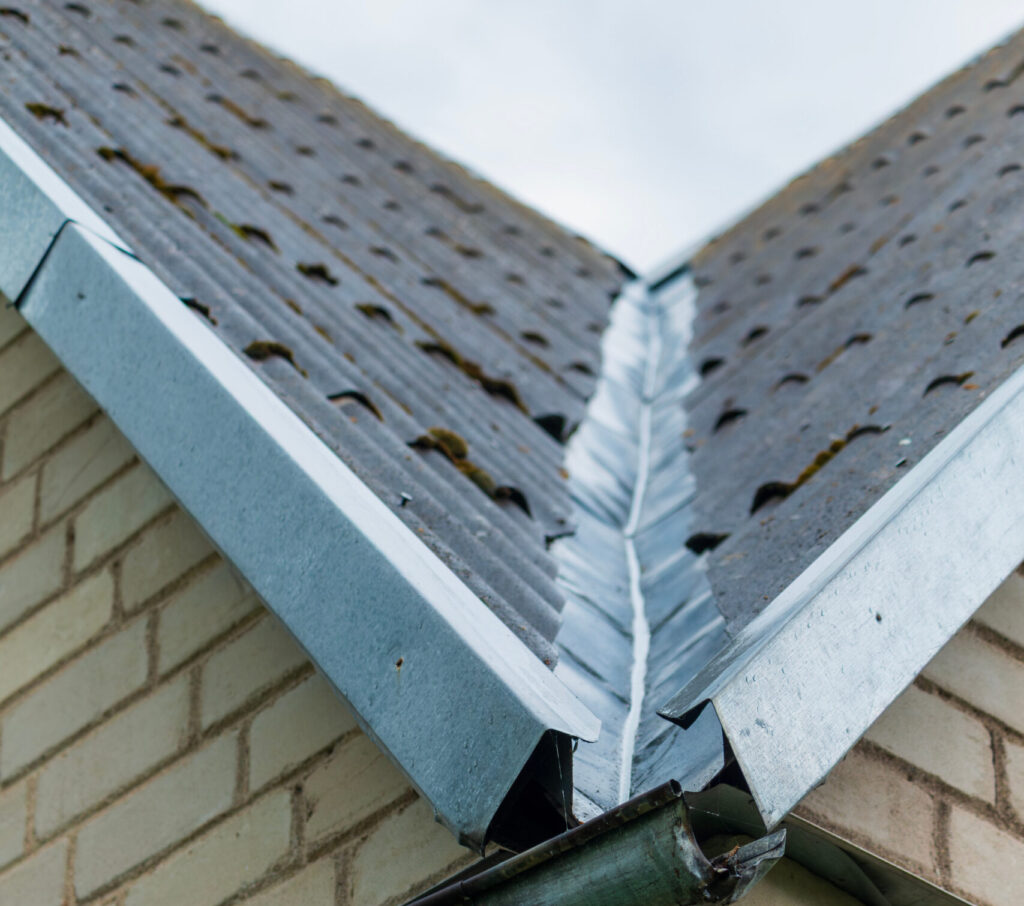 An aged residential building displays its roof, valley flashing, and rain gutter with a downspout.
