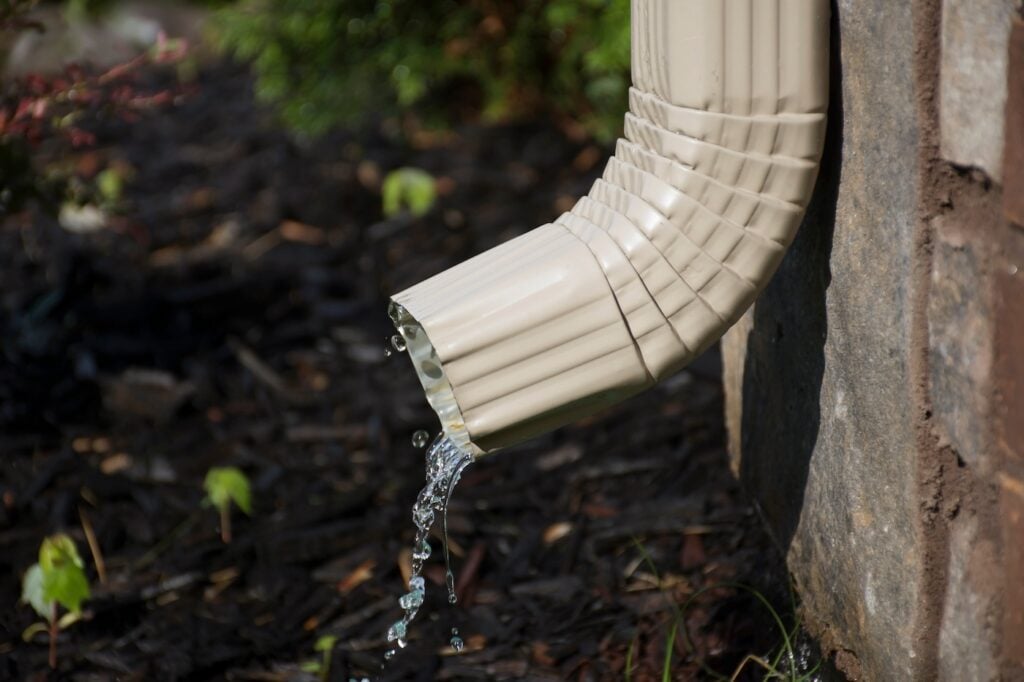 A rain gutter downspout with water coming out from the side, with a flower bed in the background