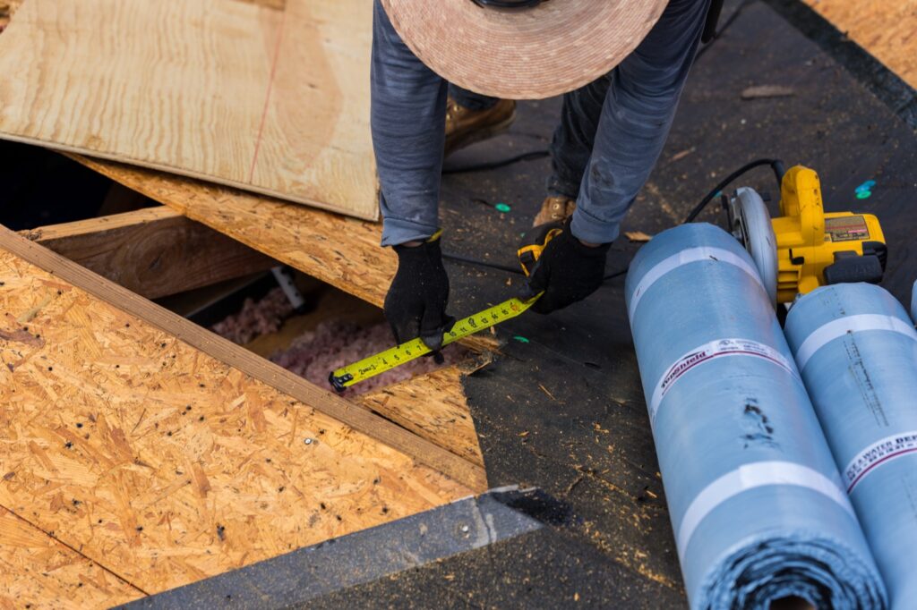Quality Roofing Solutions crew member measuring a roof deck repair on a Pensacola home, with rolls of self-adhered ice and water membrane ready to seal the deck